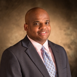 Headshot of Athan Lindsay wearing a striped shirt, a patterned tie, and a suit jacket.